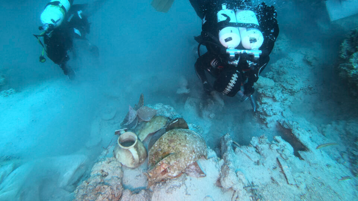 Two scuba divers explore an underwater wreck with a broken clay pot on the seabed of sand and rubble.